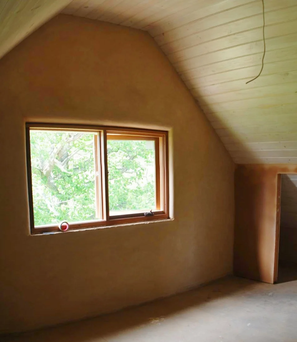 Interior showing earth-rendered walls, timber window framing and a sloping timber-lined ceiling